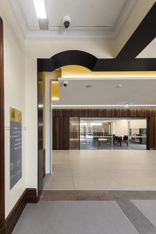 Interior view of a refined commercial building corridor or lobby, looking through a broad doorway into a bright reception or waiting area. The space features cream painted walls, dark timber skirting and trim, a dramatic black curved portal detail, a decorative white coffered and ribbed ceiling, large pale floor tiles, and a rear wall of vertical dark timber slats. Directional signage and security cameras are visible, reinforcing the public or institutional setting.