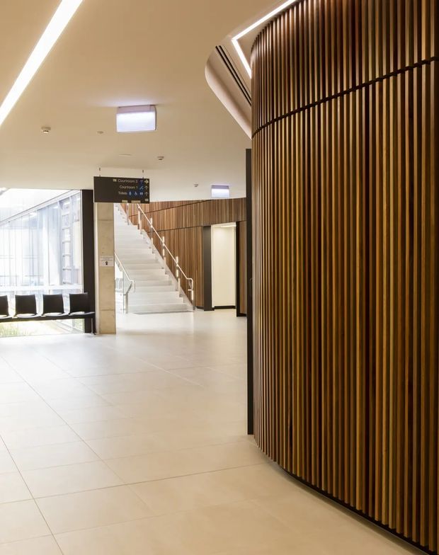 Bright contemporary institutional interior, likely a commercial lobby or public building foyer. The space features a large curved wall clad in closely spaced vertical timber slats, a pale tiled floor, white ceiling with linear lighting and skylights, a broad glazed opening, seating, and a white staircase with stainless steel handrails. Directional signage and circulation areas reinforce the lobby or public-building character.