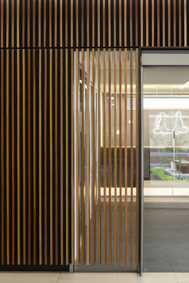 A close-up architectural view of a contemporary commercial entrance featuring a vertical timber batten screen across a dark wall and glazed doorway. The screen creates a layered privacy partition, with a bright reception or lobby visible beyond the glass, including a tiled floor, illuminated ceiling, and interior branding.
