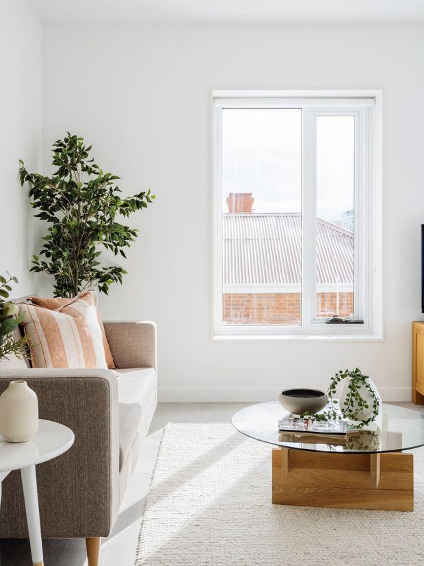 Bright, calm contemporary living room with white walls and flooring, a neutral upholstered sofa, pale textured rug, round glass coffee table with a timber pedestal base, and indoor plants. A large white-framed window provides strong natural light and views toward neighbouring rooftops. The room has a clean, airy appearance with warm wood and beige accents.