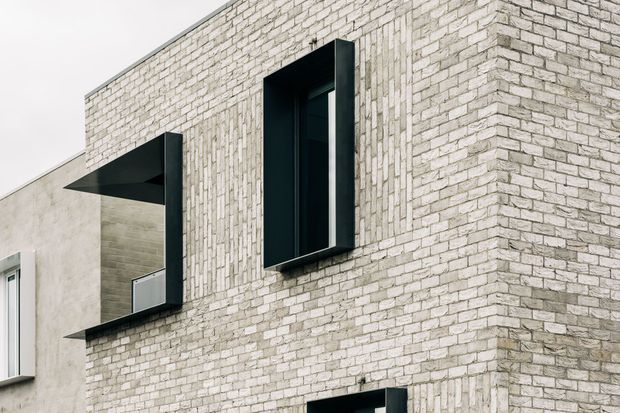 Close-up exterior view of a contemporary building facade featuring pale textured brickwork, deep-set black-framed rectangular windows, and strong projecting geometric window surrounds. The composition emphasizes the masonry wall and contrasting dark openings.
