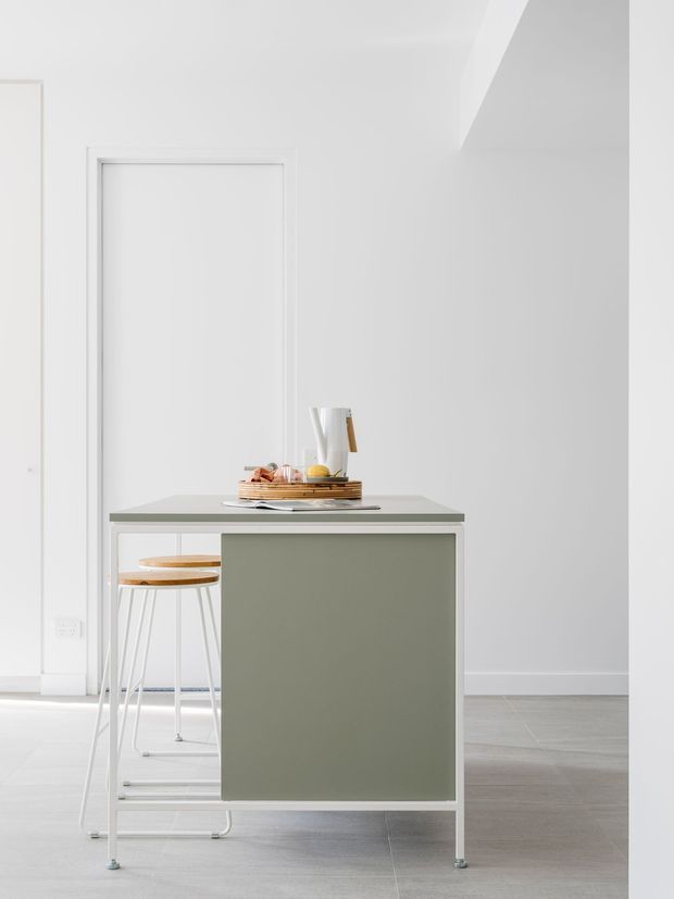 Minimalist bright interior featuring a compact kitchen island or peninsula with a muted sage-green fixed cabinet front, pale grey benchtop, and slim white legs. Two simple stools sit beneath the overhang, while a tray with crockery and a jug provides restrained styling. The surrounding space has white walls, a flush white door, pale grey tiled flooring, and clean contemporary detailing.