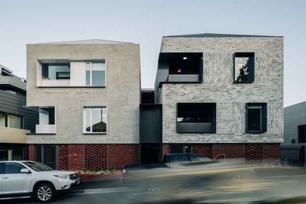 Street-level exterior photograph of a contemporary multi-storey residential development composed of two adjoining cubic buildings. The left volume has a smooth light-rendered facade over a red brick base, while the right volume is clad in pale brick with dark-framed recessed windows and a red brick lower level. A dark recessed entry link separates the volumes. Cars and motion-blurred traffic occupy the foreground.