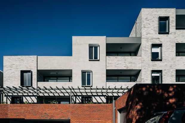Exterior architectural photograph of a contemporary multi-storey residential building, viewed from the street. The facade combines pale rendered cubic volumes with light brickwork, recessed balconies, dark-framed windows, and a linear pergola above a red-brick boundary wall. The composition emphasizes the building envelope and geometric massing against a clear blue sky.
