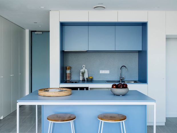 A compact contemporary kitchen with a blue island and matching blue recessed cabinetry. The island has a pale blue benchtop and white structural frame, with two timber-topped stools. Behind it is a blue galley-style cooking and sink zone featuring a small patterned mosaic splashback, integrated cabinetry, and a black curved tap. White full-height cabinetry and pale grey tiled flooring complete the clean, streamlined interior.