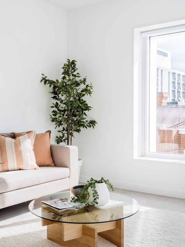 Bright, minimalist living room with white walls and flooring, a cream upholstered sofa with tan and striped cushions, a round glass coffee table on a light textured rug, and potted greenery. Large white-framed window provides abundant natural light and overlooks nearby urban buildings.