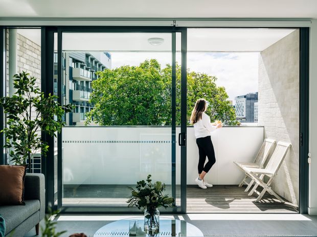 A bright contemporary apartment living room opening onto a compact balcony terrace through full-height black-framed sliding glass doors. The balcony has a white rendered privacy wall, timber-look decking, and two folding deck chairs, with leafy urban buildings visible beyond. Inside, a grey sofa, potted greenery, and a round glass coffee table sit in a calm, light-filled space.