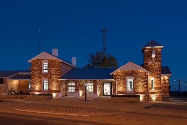 Night-time exterior view of a low-rise institutional or commercial building composed of several connected gabled sections and a prominent square tower. The facade is clad in warm irregular stone masonry with contrasting dark metal pitched roofs, white-trimmed windows and doors, and warm architectural uplighting. The building fronts a paved street or forecourt, with limited surrounding context visible.