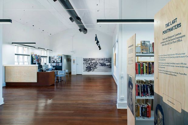 A bright, open-plan commercial interior resembling a library, gallery, or educational exhibition space. The room has polished dark timber flooring, white walls and ceiling, exposed black services ducting, linear pendant lighting, shelving filled with books, display panels, a timber reception or service counter, and a small seating area. Large windows provide natural light, while interpretive exhibition panels and a large wall graphic create an institutional cultural-space atmosphere.