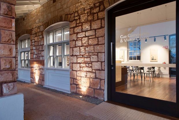 Exterior view of a heritage-style stone building entrance, with warm uplighting washing over coursed masonry and arched window surrounds. A black-framed glazed door opens into a bright visitor centre with white walls, timber flooring, and a communal table visible inside. The image combines historic architectural fabric with a contemporary public interior.