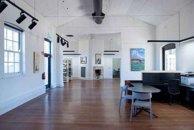 Bright, open-plan commercial interior with white walls and a high white ceiling featuring exposed ductwork and suspended black track lighting. The space has dark timber flooring, black built-in reception or work cabinetry, desks and chairs, large multi-pane windows, and framed artwork. A partially visible adjoining room and kitchenette or office area appear in the background.