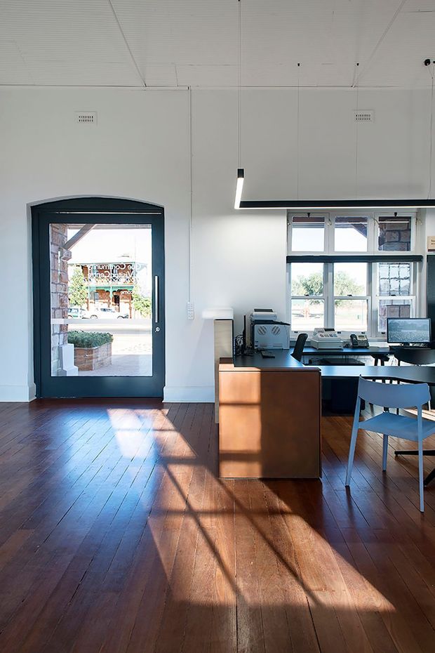 Bright, minimalist office or reception interior with a dark-framed glazed entry door, large multi-pane windows, white walls and ceiling, warm timber flooring, and a compact timber reception desk. A workstation with computer equipment, printer, and blue chairs occupies the rear of the space. Strong afternoon sunlight creates long shadows across the floor.
