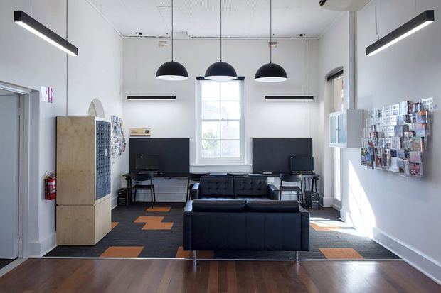 Bright, modern commercial lounge or reception area with a black leather sofa facing a pair of computer workstations. The room has white walls, polished dark timber flooring, black pendant and linear lighting, a central window, wall-mounted display racks, and a light timber storage unit. Orange geometric carpet insets add a restrained accent colour.