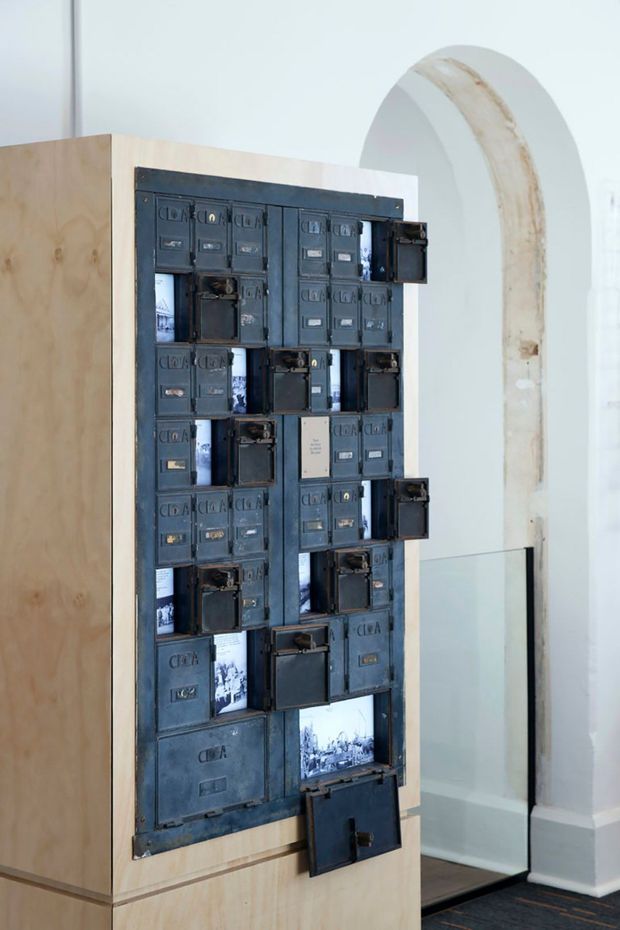 Interior architectural detail featuring a tall, freestanding pale timber display structure with an inset grid of aged dark metal compartments and hinged doors, some open to reveal black-and-white imagery. The installation stands against a bright white interior with a tall arched opening and dark flooring visible behind it. The scene has a gallery-like, adaptive-reuse character.