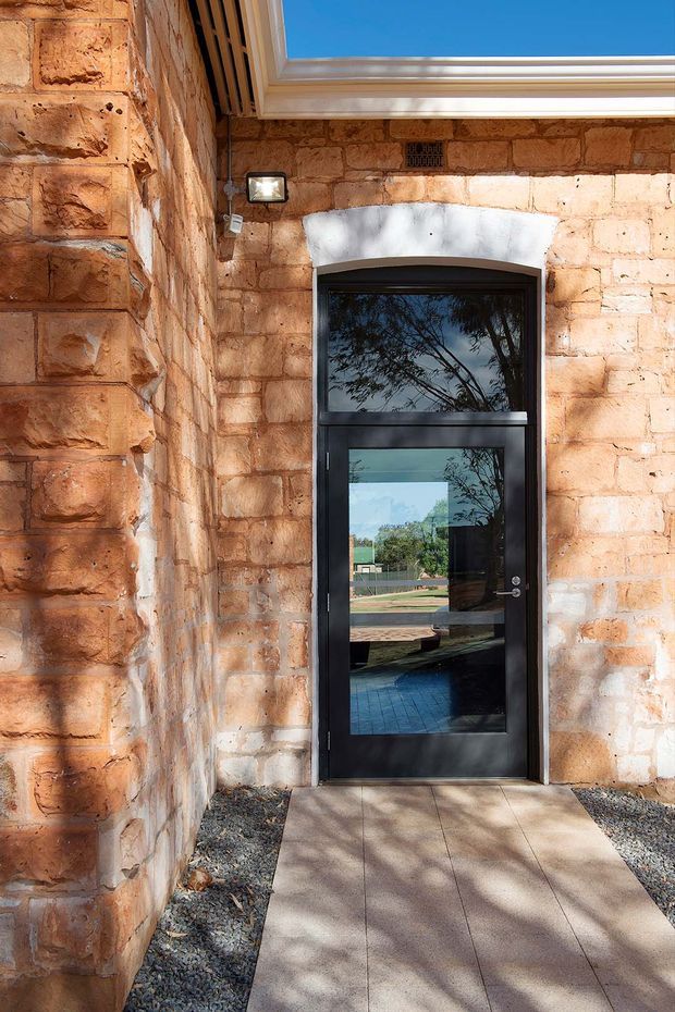 Exterior entry view of a masonry building, featuring warm sandstone block walls, a black glazed metal-framed door, and a covered or recessed walkway. A rectangular rooflight or skylight brings daylight into the entry area, while gravel borders the paved path.