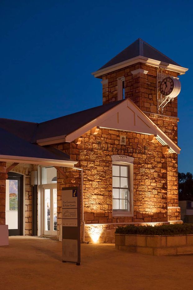 Dusk exterior view of a heritage-style stone civic or community building. The facade features warm rough-cut masonry, a steep pyramidal roof, decorative cornices, tall divided-light windows, and a prominent clock tower. Exterior uplighting highlights the stonework, while an entrance and freestanding community sign are visible at ground level.