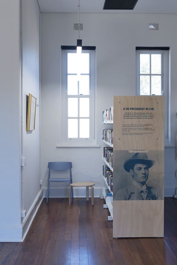 A bright, narrow heritage-style interior resembling a small library, archive, or exhibition space. White walls and tall multi-pane windows frame dark polished timber flooring. A compact blue-grey chair and round side table sit beside built-in or freestanding bookshelves, while a large timber display panel presents historical information and a portrait. The space has simple contemporary lighting and visible ceiling ventilation grilles.