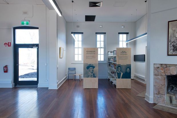 A bright, sparsely furnished heritage-style exhibition or gallery interior with white walls, polished dark timber flooring, tall multi-pane windows, suspended linear lighting, and freestanding interpretive display panels. A stone fireplace is visible on the right, with framed artwork and shelving along the walls. The space appears institutional or cultural rather than residential.