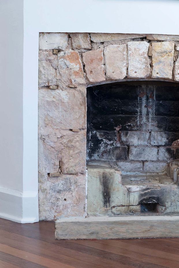 Close-up interior view of a traditional masonry fireplace with a rough, weathered stone surround, exposed brick-lined firebox, and substantial stone hearth. The fireplace is set against white plaster walls and bordered by dark timber floorboards, highlighting the aged texture and layered construction of the feature.