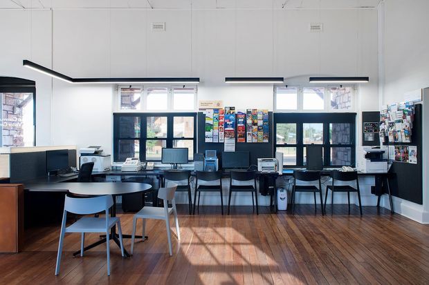 A bright, contemporary commercial interior resembling a school, library, or community workspace. The room contains multiple computer workstations, printers, shelving, display boards, tables, and chairs arranged along the perimeter and in the open central area. Large dark-framed windows and glazed doors provide strong daylight, while linear black pendant lights hang from a high white ceiling. The floor is finished in warm timber boards, creating contrast with the predominantly white walls and black-and-white furnishings.