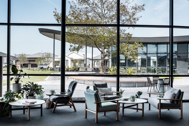 A refined contemporary lounge opening onto a landscaped courtyard and reflecting pool through expansive black-framed floor-to-ceiling glazing. The room contains several low-profile upholstered lounge chairs and sofas with timber arms, round coffee tables, indoor plants, and a dark carpet. Outside, a covered terrace, mature tree, paving, and a modern glazed building are visible.