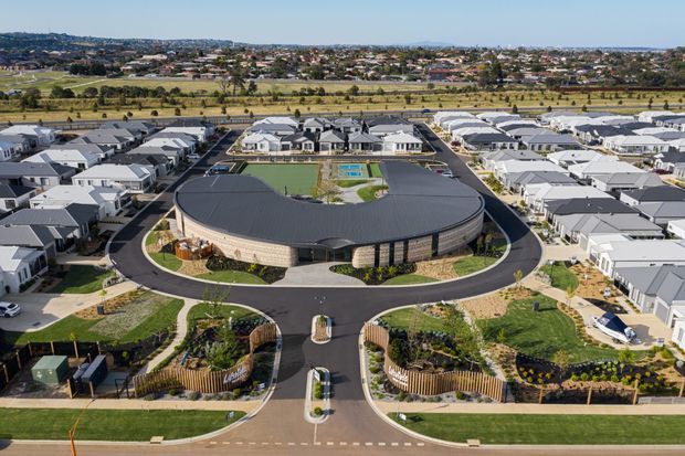 Aerial view of a contemporary oval-shaped community building or school set within a newly developed suburban neighbourhood. The building has a broad dark curved roof, pale masonry or textured facade, glazed entrances, and landscaped grounds with looping asphalt driveways, lawns, planted garden beds, and outdoor recreation areas. Rows of modern detached homes surround the site, with agricultural fields and a distant suburban landscape beyond.
