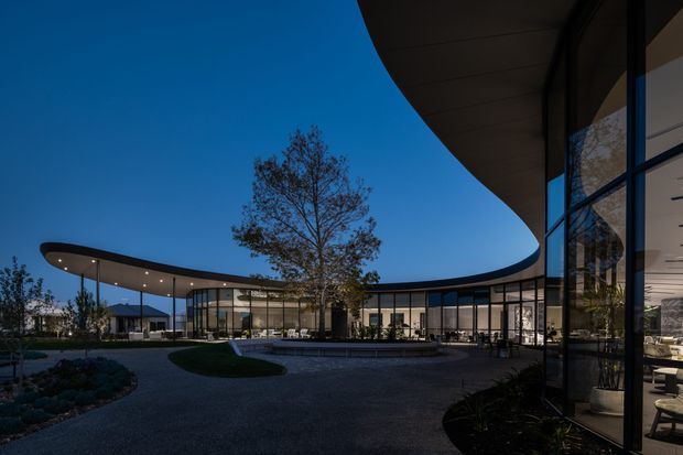 A dusk exterior view of a sophisticated low-rise building organized around a landscaped courtyard and shallow reflecting pool. Sweeping curved roof canopies extend over extensive floor-to-ceiling glazing, with warm interior lighting visible through the glass. The scene includes paved paths, planted garden beds, a mature central tree, and a dramatic contemporary architectural composition.