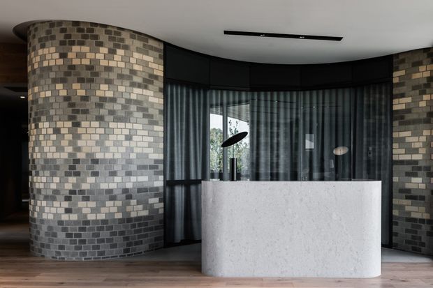 A sophisticated curved reception or kitchen-like built-in counter dominates a dark, rounded interior. The space features cylindrical walls clad in small rectangular grey, beige and cream tiles, dark-framed floor-to-ceiling glazing with sheer curtains, a smooth white stone-like counter or island, timber flooring, and a linear recessed ceiling light. The overall composition is minimalist and contemporary with a moody, refined atmosphere.