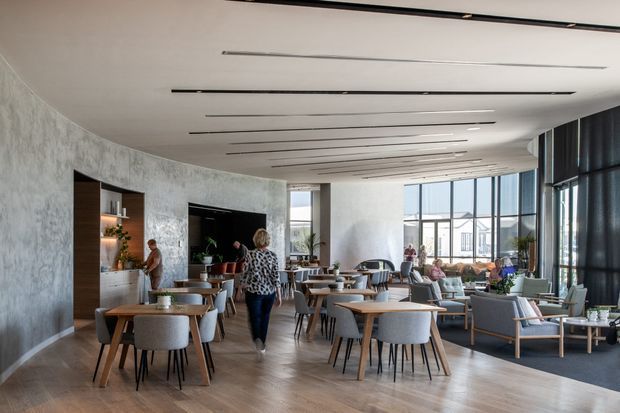 A spacious, contemporary hospitality lounge or café interior with a curved textured feature wall, integrated timber display shelving, extensive floor-to-ceiling glazing, and a softly curved ceiling. Numerous timber dining tables with upholstered chairs are arranged across a warm timber-look floor, while a relaxed seating area occupies the window side. Slim linear recessed ceiling lights create an airy, refined atmosphere.