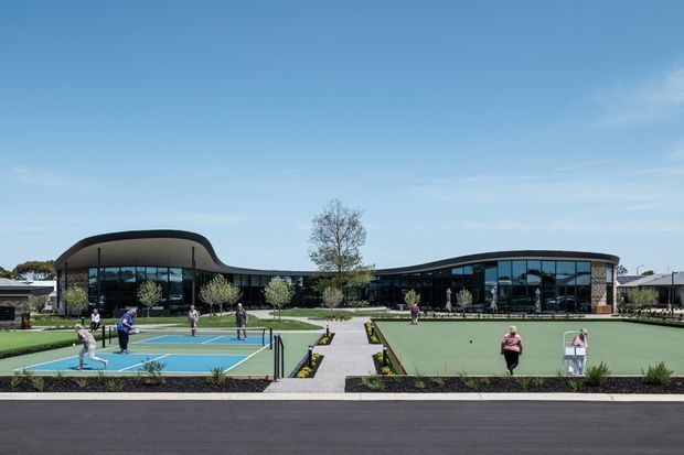 Wide exterior view of a low, sprawling contemporary complex with curved rooflines and extensive floor-to-ceiling glazing. The landscaped grounds include outdoor courts and a large green recreational area connected by paved paths, with clear blue sky and sparse planting.