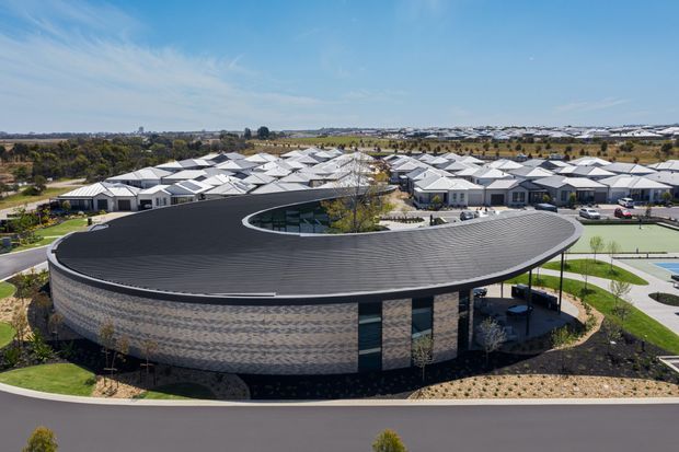 Aerial view of a contemporary curved, low-rise building with a broad dark ribbed roof and patterned masonry facade. The building is set within landscaped grounds and appears to be part of a larger planned development, with many light-roofed buildings and marked outdoor recreational courts visible behind it.