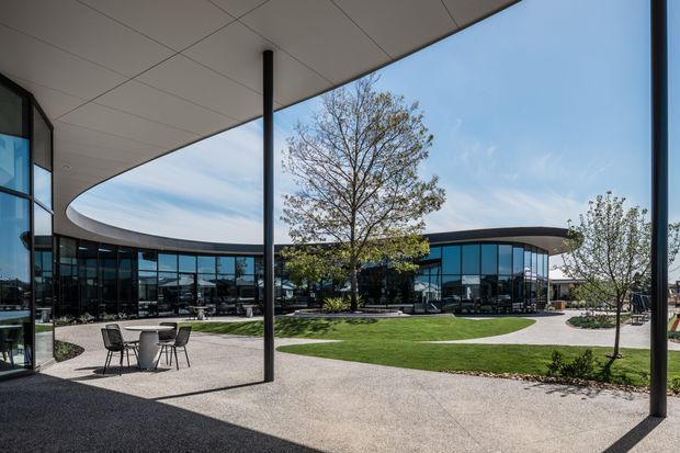 Contemporary commercial campus courtyard viewed from beneath a broad curved canopy. The scene features extensive floor-to-ceiling glazing, dark slender structural columns, a curved low roofline, paved pedestrian areas, outdoor café-style tables and chairs, and landscaped lawn with mature and young trees. The architecture has a clean, refined, open and highly glazed appearance.