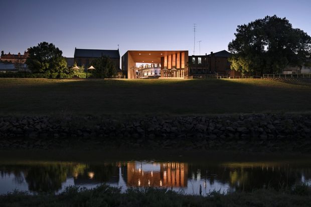 A contemporary low-rise building viewed across a grassy embankment and reflective waterway at dusk. The illuminated central entrance or pavilion has a deep flat-roofed canopy, warm timber-toned vertical fins, and extensive glazing, flanked by darker pitched-roof volumes and mature trees. The building and its lights are reflected in the water in the foreground.