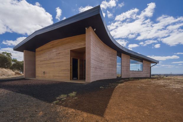 Single-storey contemporary building viewed from outside, with warm horizontal timber cladding, textured earthen or rammed-earth-like walls, expansive glazing, and a dramatic curved roof sweeping upward at the edges. It sits on a sparsely landscaped, earthy site beneath a bright partly cloudy sky.