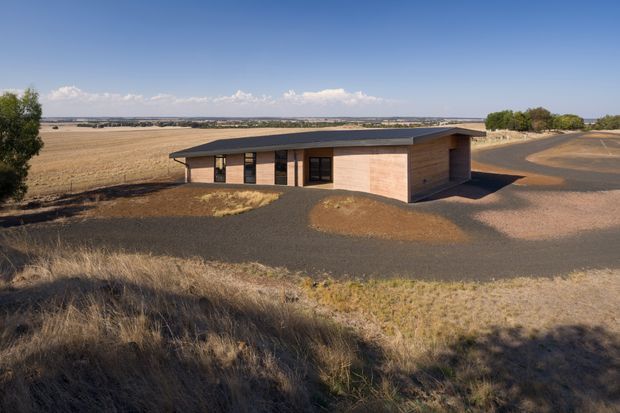 A low, single-storey modern building set within a broad, dry rural landscape. The structure has a simple elongated form, a shallow dark metal roof with generous overhangs, warm timber-clad walls, and dark-framed windows and doors. Gravel and paved access areas surround the building, with expansive agricultural fields and distant horizon beyond.