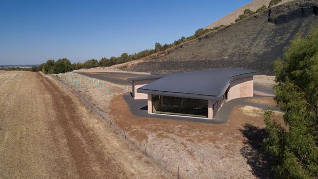 Aerial view of a low, contemporary building set into a dry, sloping rural or semi-rural site. The structure has a broad dark curved roof, pale masonry or rendered walls, extensive glazed frontage, and a driveway or access path wrapping around it. Cultivated rows, sparse vegetation, exposed earth, and a steep cut hillside surround the building.