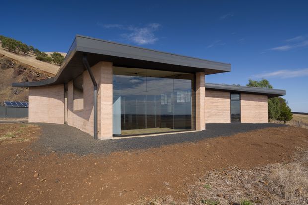Exterior view of a low-slung contemporary building set on a dry, gently sloping rural site. The structure features broad flat roof planes with deep overhangs, exposed dark structural supports, warm horizontal masonry walls, and a large floor-to-ceiling glazed entrance or living area. Gravel surrounds the building, with sparse vegetation and open countryside in the background.