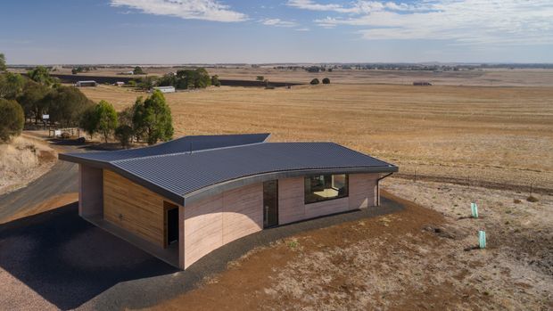 Aerial exterior view of a compact single-storey contemporary building set within expansive harvested agricultural fields. The structure has a low-pitched dark metal roof, timber-clad walls, large glazed openings, and a simple rectangular form, with a paved driveway and sparse landscaping around it.