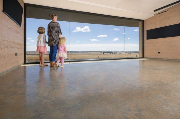 A spacious, minimally furnished living area with a polished concrete floor, exposed textured concrete walls, and expansive floor-to-ceiling glazing overlooking a broad rural landscape. A woman and two children stand near the window, while dark wall-mounted rectangular features and a slim linear ceiling vent add contrast. The room has a contemporary, industrial character and abundant natural light.