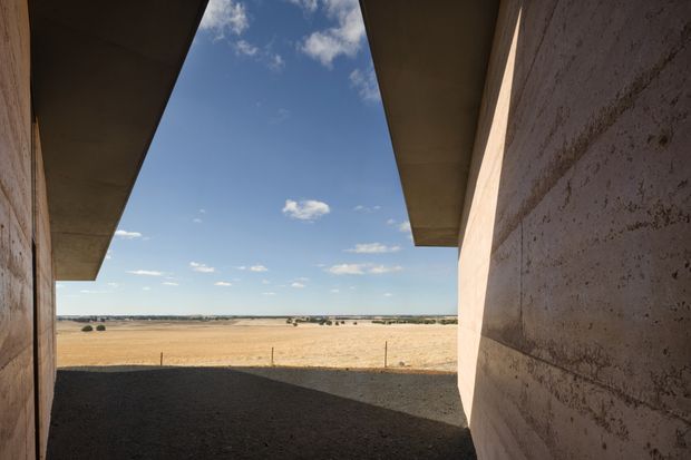 A view from a narrow, shaded architectural passage or courtyard opening toward a broad rural agricultural landscape. The structure has massive raw textured concrete walls and deep projecting flat roof slabs, creating a strongly framed vista of dry golden fields beneath a clear blue sky.