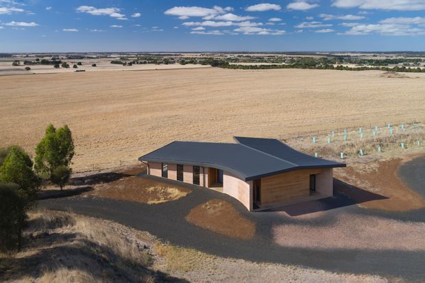 Aerial view of a low, contemporary single-storey building set within a broad agricultural landscape. The building has a dark, low-pitched hipped roof, simple timber-clad walls, extensive glazing along one side, and a dark paved or gravel forecourt. Open harvested fields and rows of young plantings surround the property.