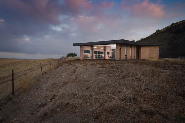 A low, minimalist single-storey house set on an exposed sloping rural or coastal landscape. The building has a broad flat roof, dark slender roof edges, extensive floor-to-ceiling glazing, and warm brown brick walls. Interior furniture is visible through the glazed central living area, while the surrounding dry grassland and rocky slope create a rugged, dramatic setting beneath a cloudy dusk sky.