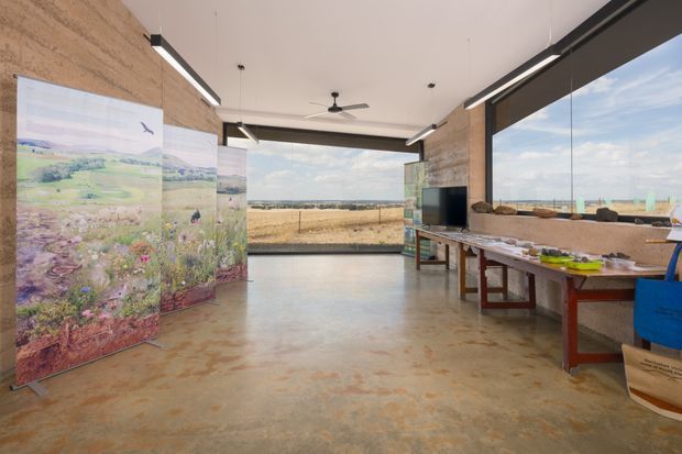 An open-plan contemporary exhibition or commercial interior with expansive floor-to-ceiling glazing overlooking a rural landscape. The space features polished concrete flooring, raw textured concrete walls, linear suspended lighting, a ceiling fan, a television, and long work or display tables. Large illustrated panels create a temporary exhibition installation, while the restrained industrial material palette and panoramic glazing give the room an airy, functional character.