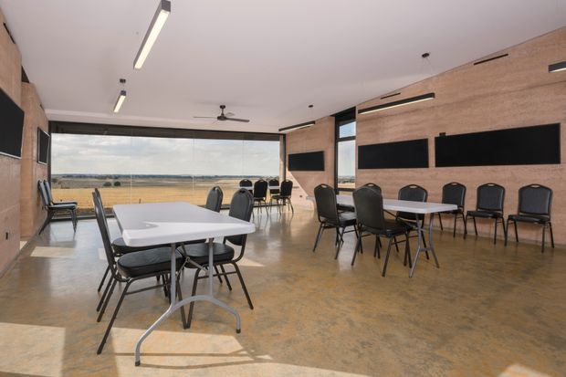 A bright, spacious multipurpose meeting or dining room with rows of tables and black upholstered chairs. The room has warm timber wall panelling, polished concrete flooring, large full-height glazed openings, wall-mounted black display panels, linear ceiling lights, and a ceiling fan. A flat rural landscape is visible beyond the glazing.