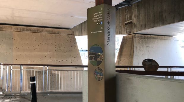 Interior view of a concrete architectural structure with exposed rough concrete walls and ceiling, metal safety railings, and an informational wayfinding pillar labelled “Mangrove Walk.” The space appears to be a covered public walkway or viewing area, with daylight visible through openings.