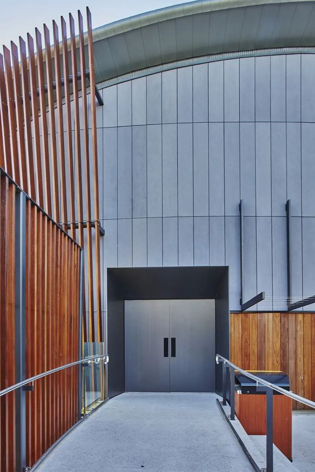 Exterior architectural view of a contemporary building entrance approached by a concrete access ramp. The facade combines pale grey modular panels, warm vertical timber screening, dark metal trim, stainless-steel handrails, and a curved standing-seam metal roof edge. Large plain double doors sit beneath a deep dark recessed canopy, with a small timber-clad service or reception element to the right.