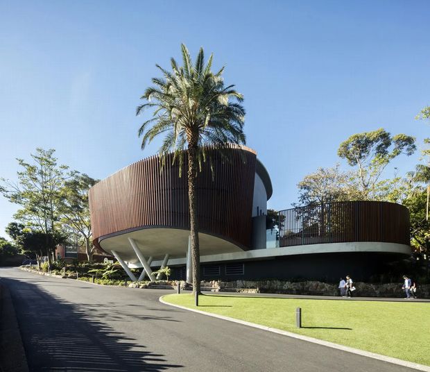 Exterior architectural photograph of a contemporary multi-level building set within landscaped grounds. The structure features a rounded, cantilevered form, dark vertical timber-like screening, extensive glazing, slender angled supports, and a curved roof terrace or balcony. A palm tree, lawn, paved driveway, and surrounding mature landscaping are visible.