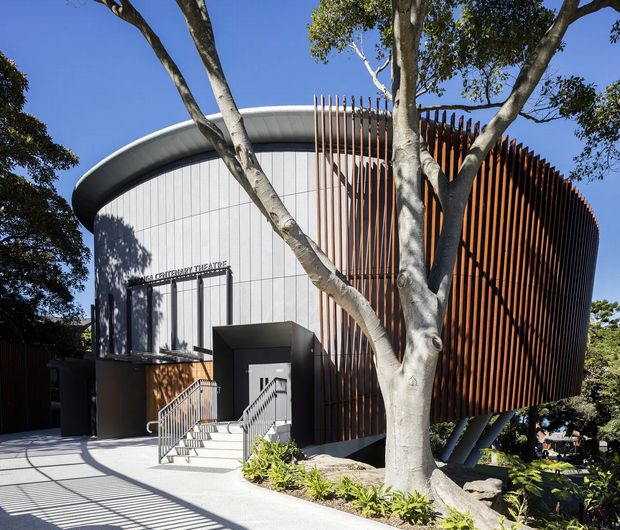 Exterior view of a contemporary performing arts theatre, with a curved white metal-clad volume and a dramatic rounded facade screened by closely spaced vertical timber fins. The entrance is reached by stairs and ramps, while mature trees frame the building and cast strong shadows across the pale paving.