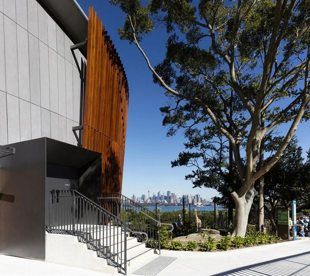 Exterior view of a contemporary building beside a landscaped pedestrian path, featuring a tall white panelled facade, dramatic vertical timber screening, dark metal entry canopy, and metal-railed stairs. A harbour and dense city skyline are visible beyond mature trees, indicating an urban waterfront setting.