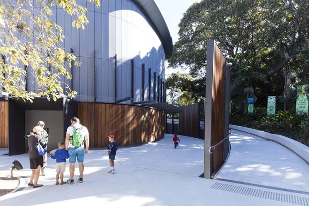 Exterior view of a contemporary curved-roof public or commercial building entrance, with dark vertical metal cladding, warm timber screening, a tall timber-and-metal gateway, and a broad pale concrete approach through landscaped grounds. Visitors and children are present near the entry.